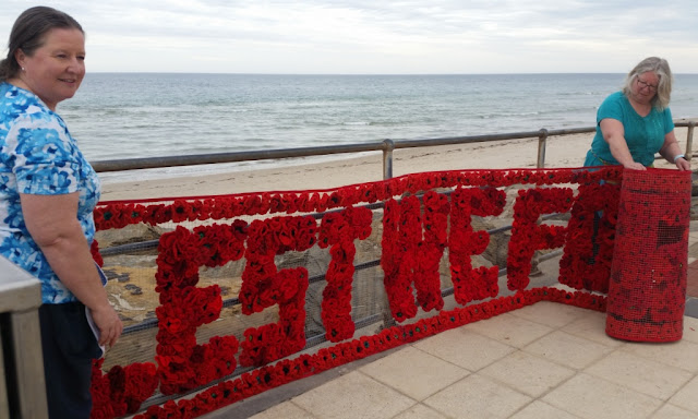 Two ladies are unrolling the "Lest we forget" banner.  The banner itself is transparent mesh and the words are made of poppies.  At left is the creator of the artwork, Dianne, holding the left hand edge of the banner while her sister, Narelle, is unrolling the other end of the banner.  Behind them are metal fencing rails, and beyond that the rocks, the beach and the waters of St Vincents Gulf. Dianne has long, dark, straight hair worn in a low ponytail and is wearing a blue and white patterned top. Narelle has shoulder length, light, straight hair and is wearing glasses and a teal top.