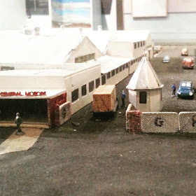Model of the entrance to a factory complex. On the veranda to the left is a sign saying 'General Motors' and a truck is going through the gate.