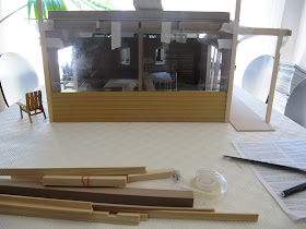 Dry fit of a dolls' house shed kit, with perspex and weatherboarding taped to the front and pergola posts and struts taped to the sides.