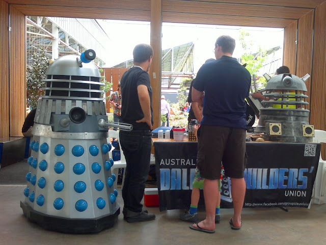 The stand for the Australian Dalek Builders' Union. Two daleks can be seen, a blue and a yellow together with visitors around the stand.