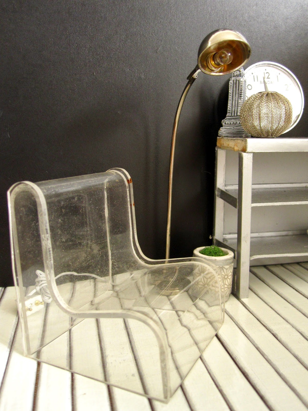 Corner of a modern dolls' house miniature interiors shop, with a clear perspex lounge chair, a silver floor lamp and a grey industrial bench on display.