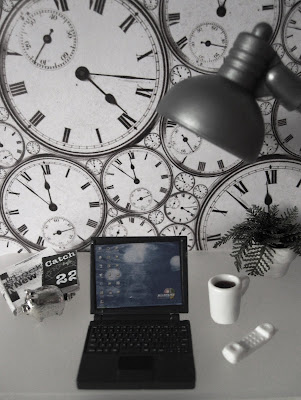 One twelfth scale modern miniature laptop computer on a desk with a phone handset, a mug of coffee, a piggy bank and two books: The shock of the new and Catch 22. The wall behind the desk is papered with a design of large clock faces.