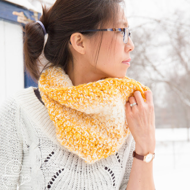 Image of a tan skin bespectacled Asian woman with dark brown hair up in a looped pony tail, looking to her left while holding the top edge of a yellow and cream crocheted cowl that's worn over a white sweater. She's in front of a worn white shed in the snow with bare branched trees in the background.