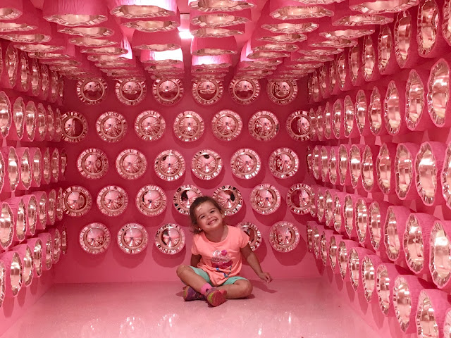 Toddler girl sitting and smiling in a pink room with chrome objects pasted to the walls and short ceiling