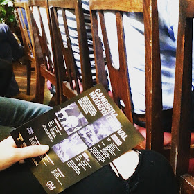 Person holding a flier for the Canberra Modern festival while seated behind a row of chairs designed by Fred Ward.