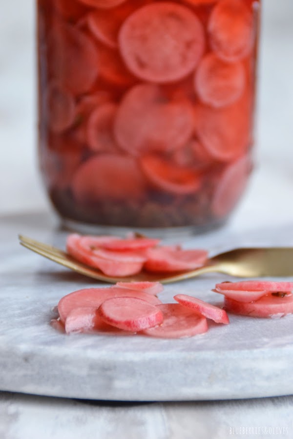 TARRO DE CRISTAL CON RABANOS MARINADOS, TENEDOR CON TROZOS SOBRE TABLA DE MÁRMOL