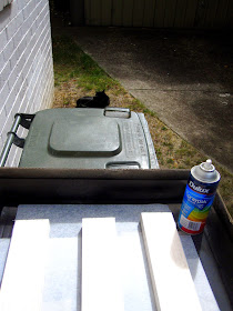 Can of spray paint and three lengths of balsa wood on top of a recycling bin outside a brick house.
