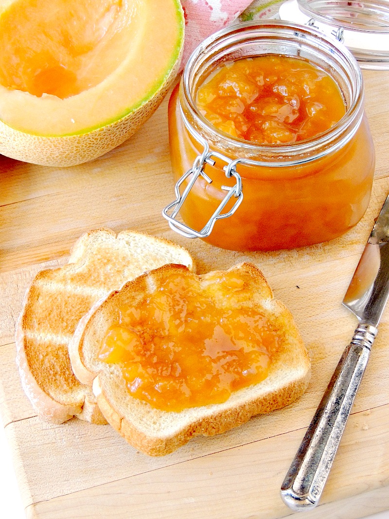 Cantaloupe Jelly in a glass jar with a fresh cantaloupe half and toast beside it on a wooden cutting board with a floral towel.