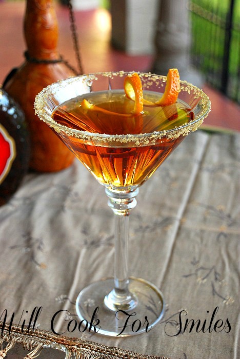 The Amaretto Peach Martini is presented in a tall martini glass. It's rim is covered in brown sugar. The liquor is gold in color. There is a swirled orange peel inside of the glass as well.
