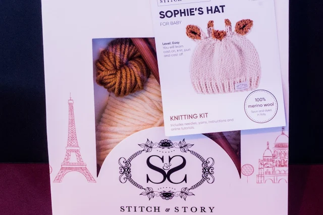 A gift package with yarn and needles visible through a window and a label with a giraffe hat based on Sophie La Girafe
