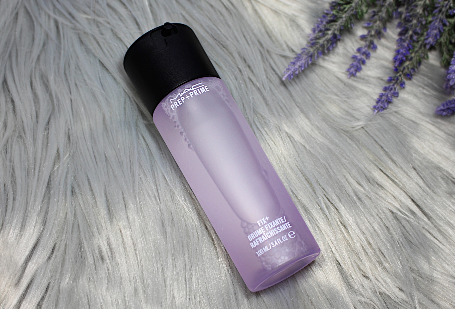 a closed bottle of Lavender Setting Mist by Mac Cosmetics is laying on a faux fur rug together with a branch of lavender flowers