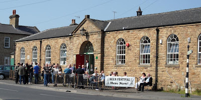 A long low stone built buiding with huge arched windows set either side of a central door, people are outside on the verge drinking and there is a banner advertising the beer festival hung on the pedestrian barriers
