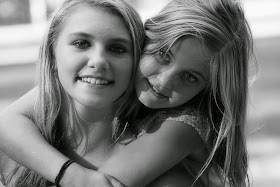 Portraitfoto: zwei junge Frauen lächeln in die Kamera, während eine die Andere von hinten umarmt.