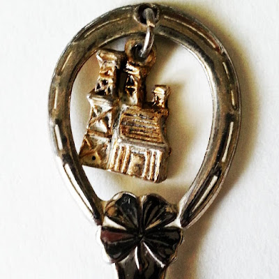 Close up of the top of a vintage souvenir teaspoon with a small metal building hanging from a hole in it.