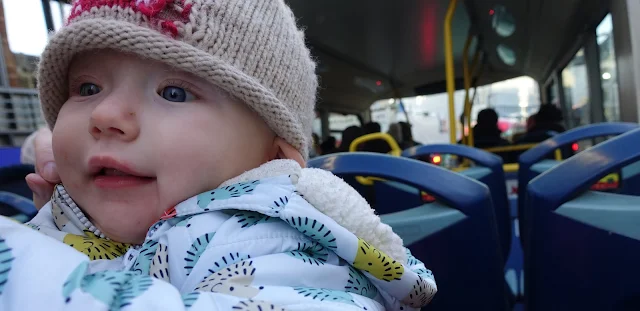 A baby in a warm hedgehog coat and woolly robin hat on the top deck of an open top bus