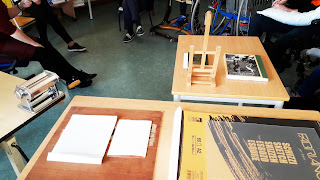 nas mesas encontram-se várias ferramentas: vários tipos de pepel, livros, cavalete pequeno, máquina de fazer massa