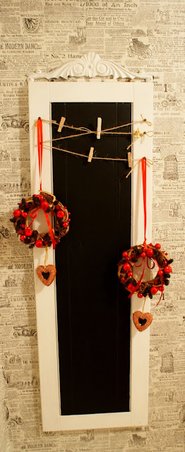 dwa małe wianuszki świąteczne na czerwonej wstążce, zawieszone na czarnej tablicy w białej ramie