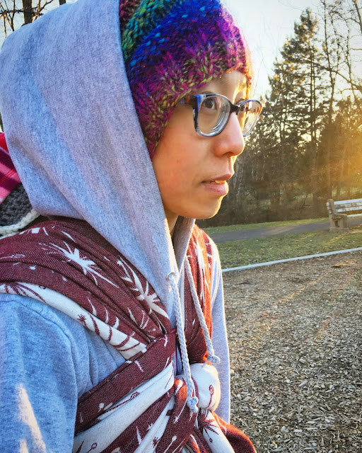 [Image of the side profile of a tan skin bespectacled Asian woman wearing a knit jewel-toned had underneath a gray hoodie from her sweatshirt. She is wearing a sleeping toddler on her back in a maroon and ecru star burst patterned woven wrap. In the background are woodchips from the playground, stately evergreen trees, a wooden park bench, and the setting autumn sun.]