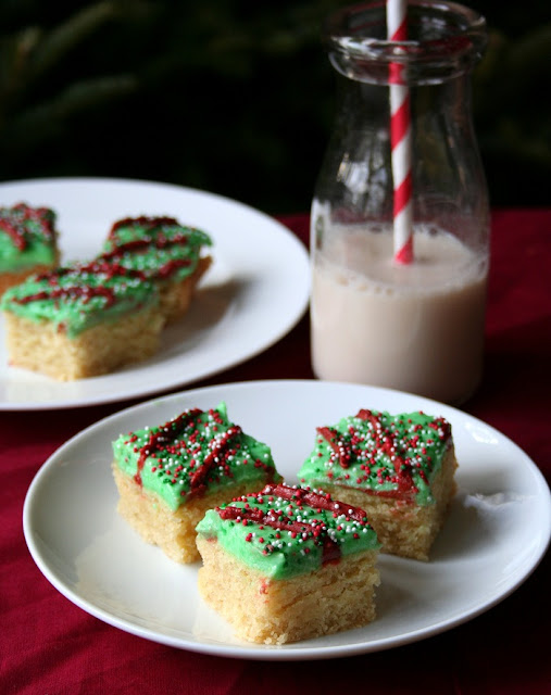 Sugar Cookie Bars – Low Carb and #glutenfree from All Day I Dream About Food inkatrinaskitchen.com #BringtheCOOKIES close up of three low carb and gluten free sugar cookie bars with green frosting and nonpareil sprinkles on a white plate and red tablecloth and more milk and cookies in the background
