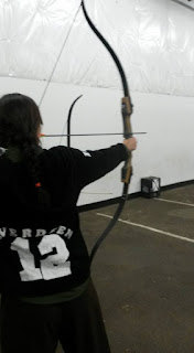 the back of a female presenting person with long braided dark brown hair. She is pulling an arrow on a left handed recurve bow and her back is to the camera. her jacket is black and says EVERDEEN on it, letter jacket style. The letters are in an arc above a large number 12