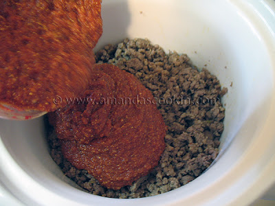 A close up photo of pizza sauce being poured over browned meat in a slow cooker. 