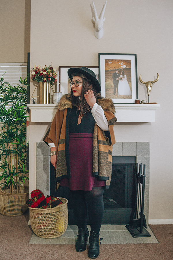 An outfit of a black wide brim fedora, brown faux fur collar poncho, peter pan collar blouse, black sleeveless dress, purple chevron print skater skirt, black tights and black heeled buckle boots.