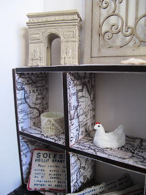 Close-up of one end of a modern dolls' house miniature pigeon hole bookcase, covered in paper printed with a vintage map of France. Displayed on it are a model Arc de triomphe, cushions with french writing on them and a chicken ornament.