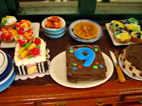 Selection of miniature dolls' house cakes arranged on a desk, including one with the number 9 iced on the top of it.