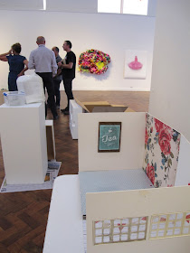 Dolls' house miniature scene on a table in a gallery during install, with gallery staff talking in the background.