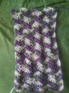 A rectangular piece of crochet lace, pooling with black, purple, and smaller patches of pale blue and white.  There is a live stitch on a wooden hook at the top of the rectangle, with a row still in progress.
