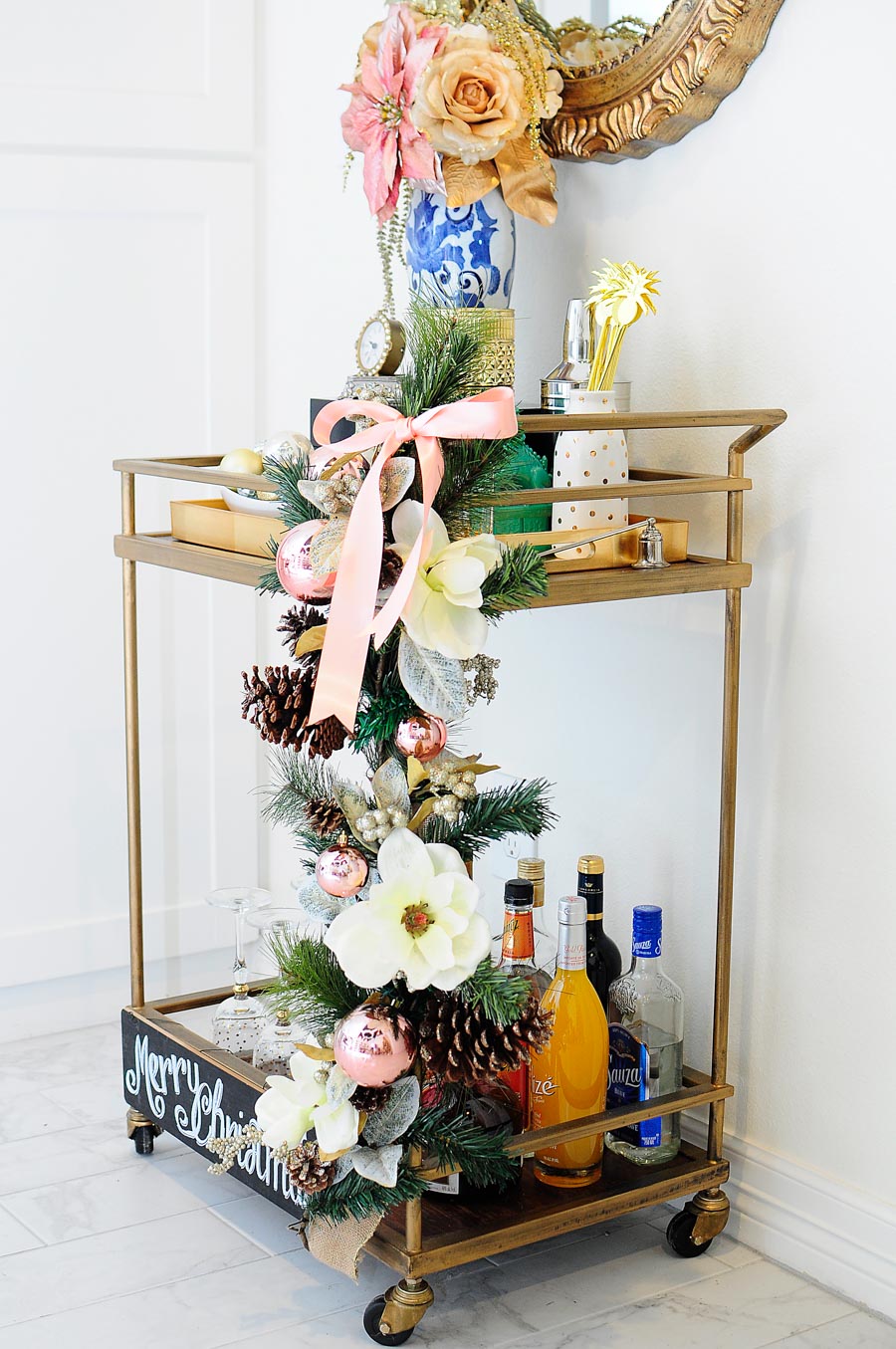 A gorgeous, eclectic bar cart decorated with blush, silver, gold and white accents, a ginger jar vase, foo dog and more. The gold mirror above the cart is perfection.