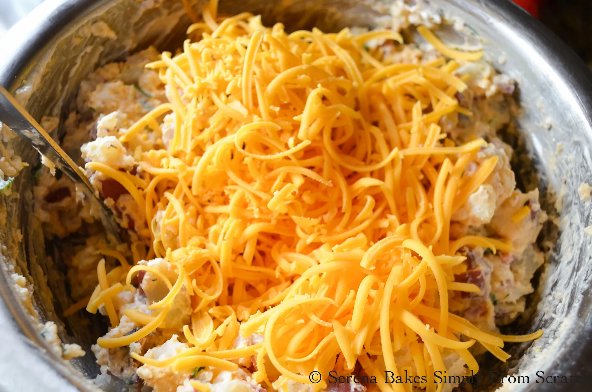 Shredded Cheddar Cheese added to Baked Potato Salad in a stainless steel bowl.