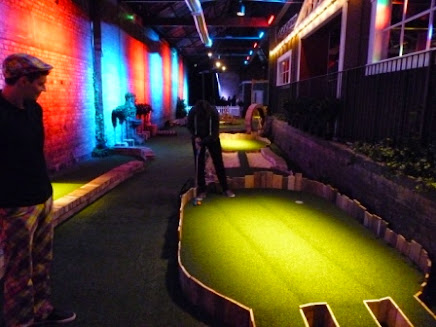 Emily playing a shot on hole 2 as one of the course guides/umpires/caddies looks-on. This hole has a tiered green and three routes to the hole Swingers Crazy Golf in Shoreditch, London