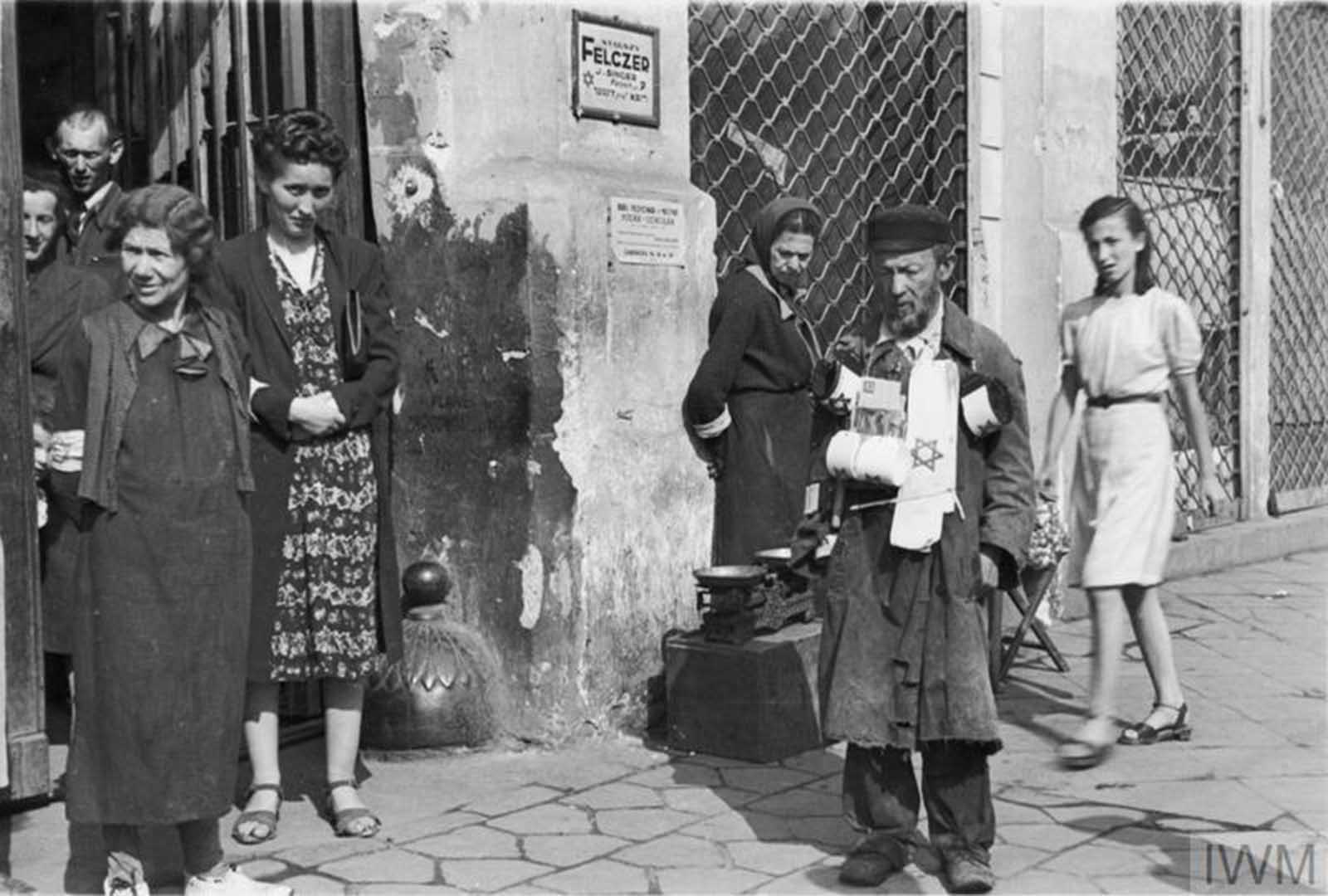 A street armband seller and a group of pedestrians on 18 Zamenhofa Street (probably) in the ghetto, summer 1941. Note two advertising plaques on the wall in the background - for Senior Medic (starszy felczer) named J. Singer and for typewriting services (address given - 18 Zamenhofa Street, flat no. 38).