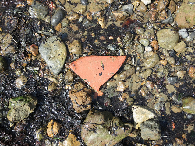 Triangular fragment of terracotta tile or pot washed up on beach with pebbles, sand and seaweed.