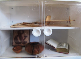 Modern dolls' house miniature glass-fronted display cupboard containing a modern wooden tray, an ampersand tile, two pieces of wood turning, and a book safe.