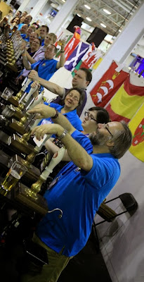 Row of blue shirted people working behind a CAMRA bar - leaning on handpumps