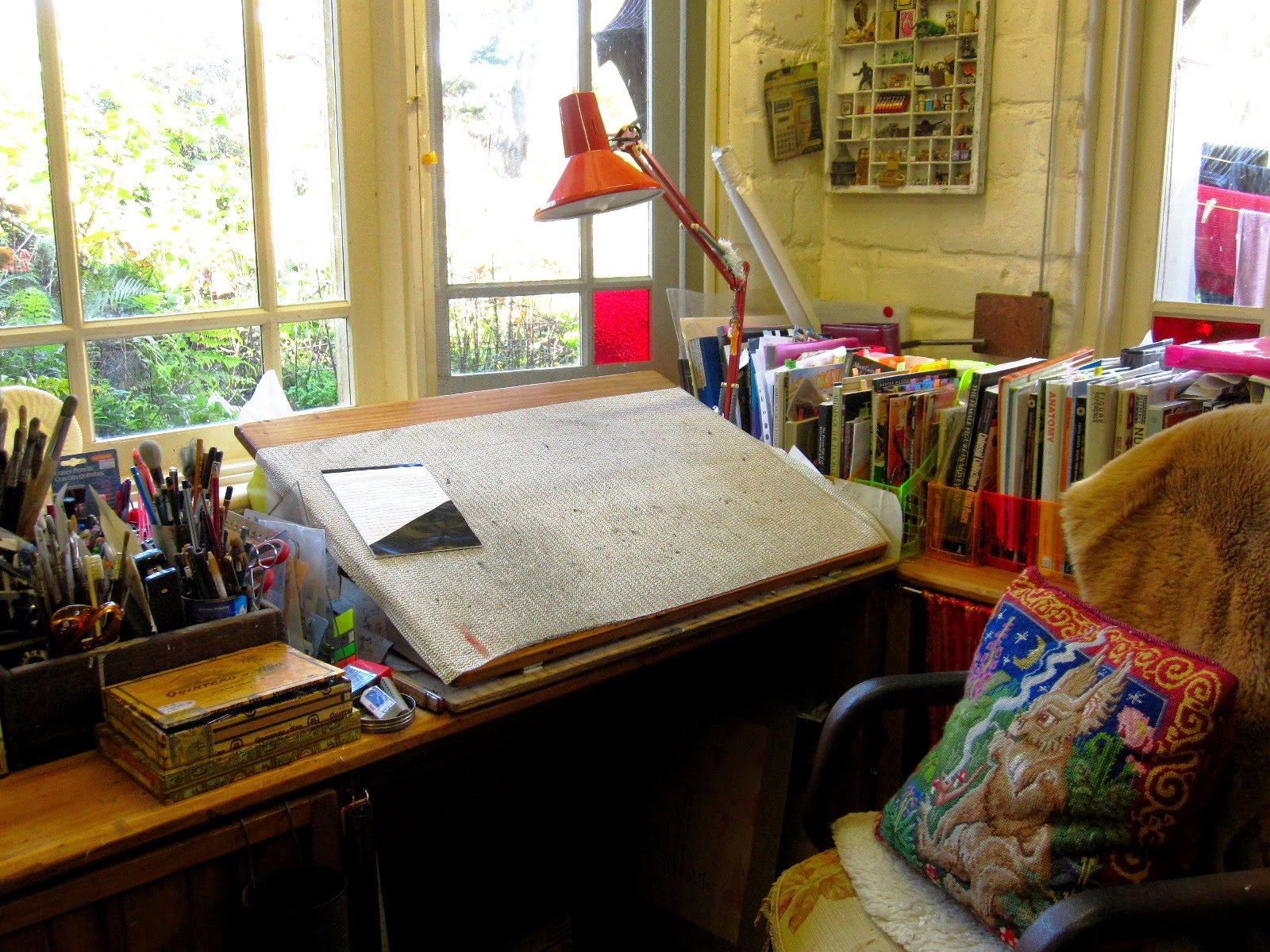 Drawing table with lamp, in the corner of a room overlooking the garden. On the desk chair is a tapestry cushion of a fantasy creature.
