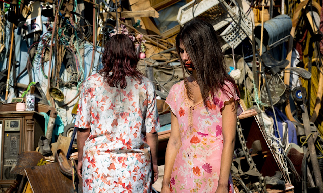 Zombie girl and her mother stand in front of a pile of junk
