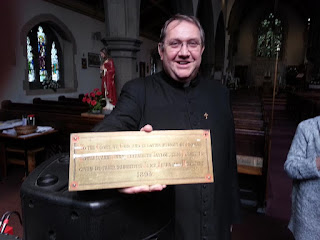 Father David in clerical dress holding out a brass plaque.
