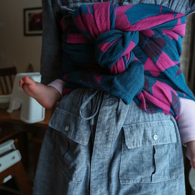 [Image of a woman's torso featuring a pink an blue highly textured woven wrap carrier in a back carry with a twist down the front and ends tucked here and there. Toddler legs dance unevenly at her sides. In the background is a breakfast table with a step stool on top of it because, children shenanigans.]