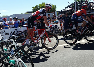 Amidst the support vehicles, two cyclists ride from left to right in the photo. On the left, the Bora team bikes are resting, in the distance are onlookers, on the right, bikes are stacked on the roof of a support car.