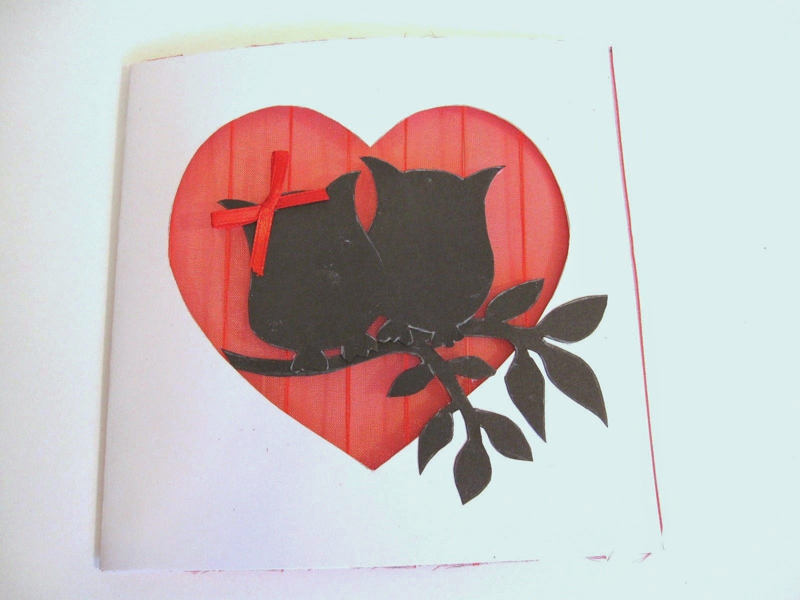 tarjeta blanca de felicitación con corazón calado con fondo de gasa roja y siluetas delante de dos búhos y rama de árbol