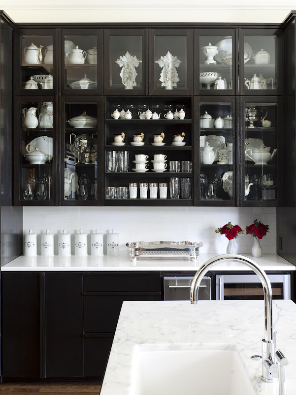 Kitchen with black lower cabinets and black upper cabinets with glass doors, white backsplash with matching counter tops and an island with a marble counter top and farmhouse sink