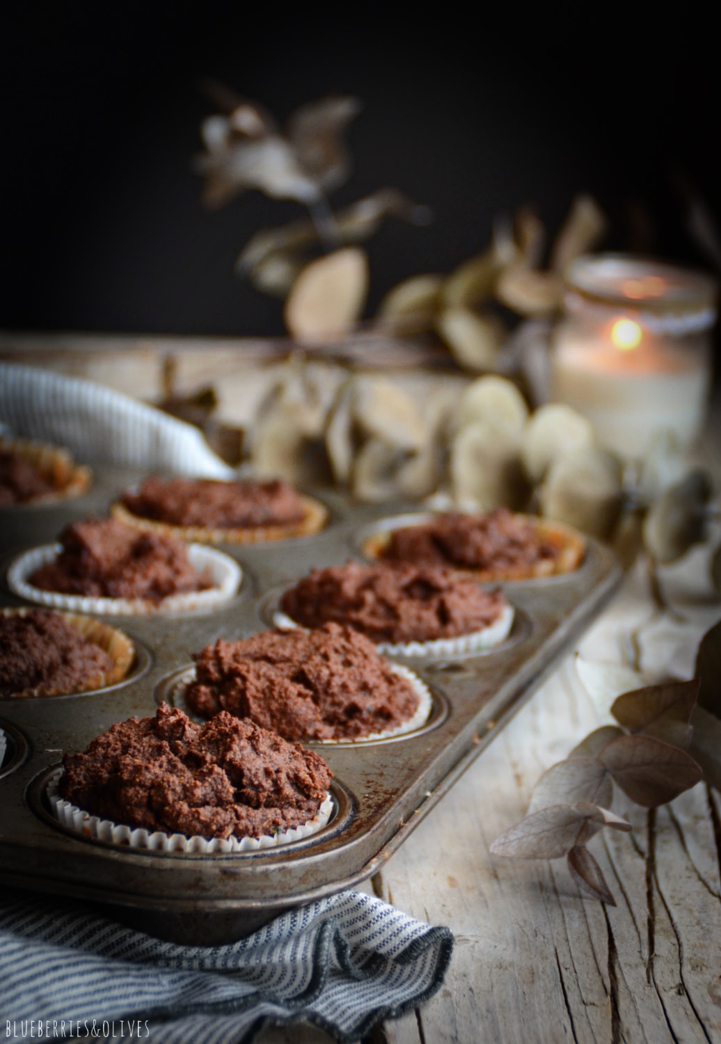 Cocoa muffins in old retro metallic pan, over old wood table, dark background, dry eucalyptus leaves, linen grey kitchen cloth
