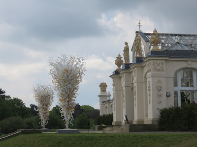 photo-  on left, Chihuly glass sculpture-  2 inverted conesmade up of white and yellow glass tentacles, on right, neo-classical white building (the temperate house at Kew Gardens)