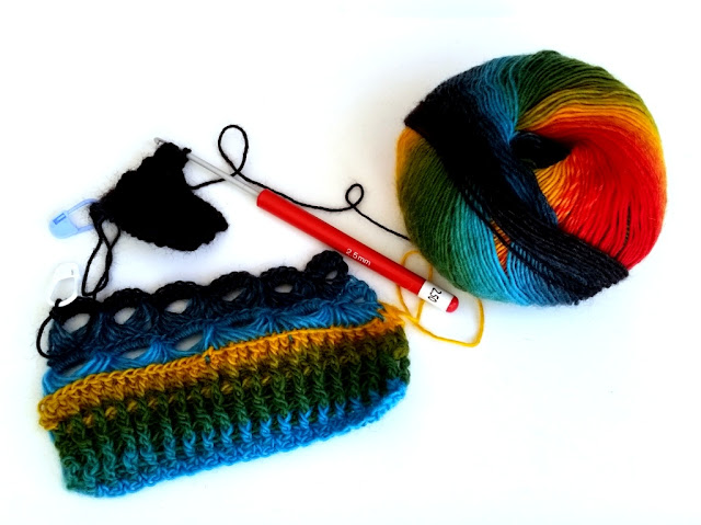 A ball of rainbow coloured yarn on the right hand side with two small samples linked to it by a dark section of yarn. The largest sample starts with light blue to green to yellow horizontal striped ribbing above which are two rows of broomstick eyelets in light blue and dark blue.  Following on from that sample, attached by a length of dark yarn, a sample of darkest blue post stitch ribbing. The crochet hook is still inserted into the active loop at the top of the dark sample. The hook is powdercoated aluminium with a red plastic handle. There is a white label attached with the size '2.50 mm' printed on the label. The size '2.5 mm' is also embossed on the side of the red handle in fine white lettering.