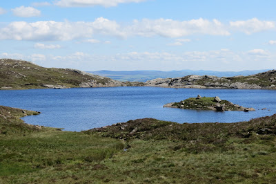 A small, blue lake with a tiny island, surrounded by moorland vegetation.