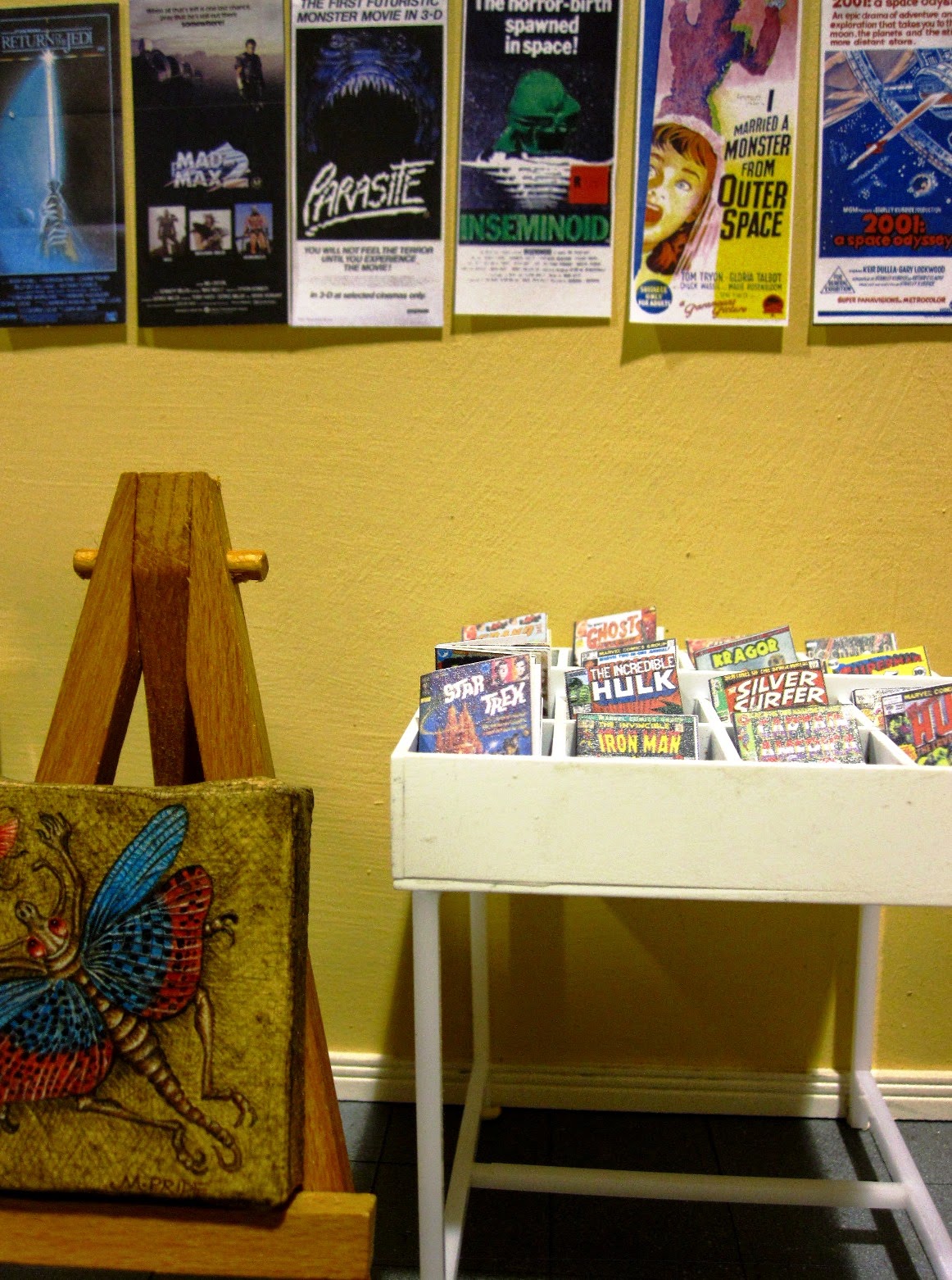 Modern miniature comic book store, interior view showing a rack of comics, a wall of posters and a fantasy art piece on an easel.