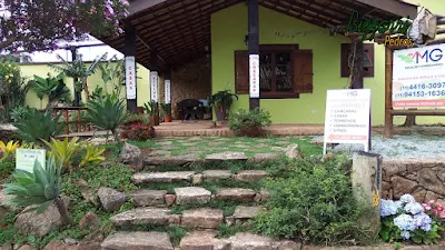 Piso com tijolo de demolição no terraço com os pilares de dormente na construção da cabana rústica com a escada de pedra, piso de pedra com junta de grama e no fundo a parede de pedra no terraço da cabana.